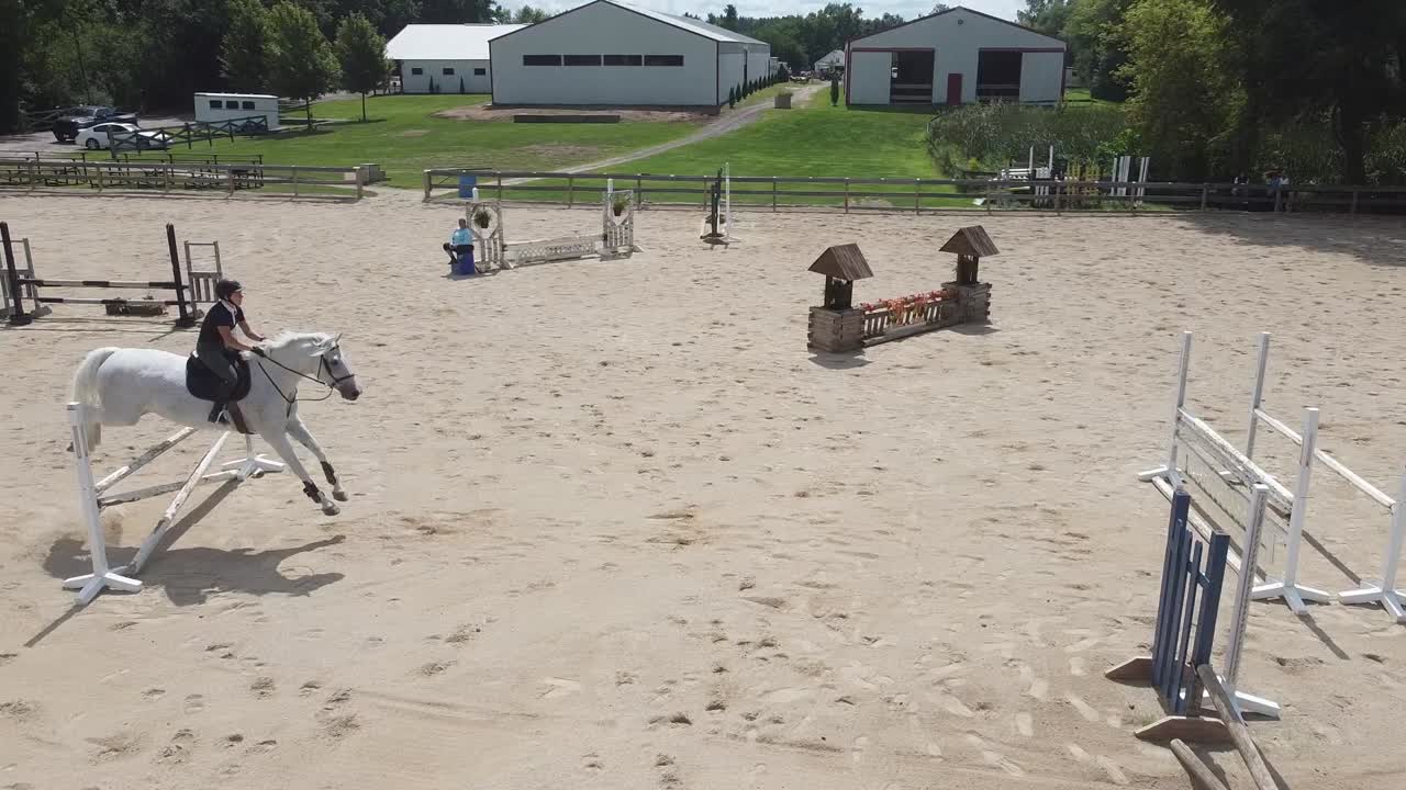 Uma imagem de drone de uma arena ao ar livre e um celeiro enquanto uma amazona e seu cavalo saltam sobre obstáculos.
