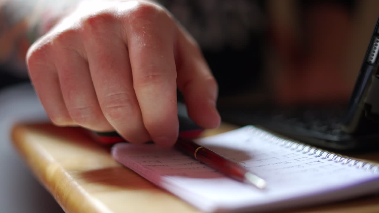Caucasian man with tattooed arm reaching for pen sitting on pad of paper and moving it out of the way of To Do list, then typing on iPad computer keyboard