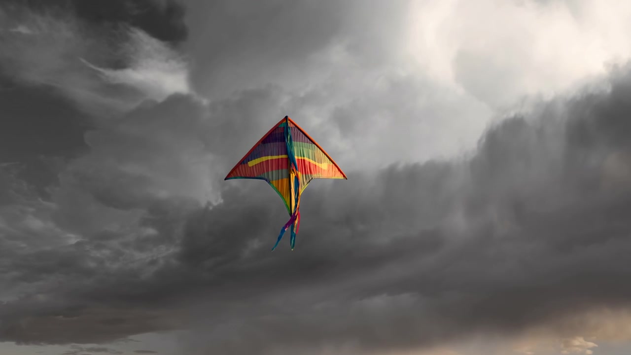 Lys, flerfarget deltakite svever høyt på en vid himmel, og viser røde, oransje, gule, grønne, blå og lilla striper, en avsmalnende trekantet form, stofftekstur og stemningsfulle stormskyer rundt.