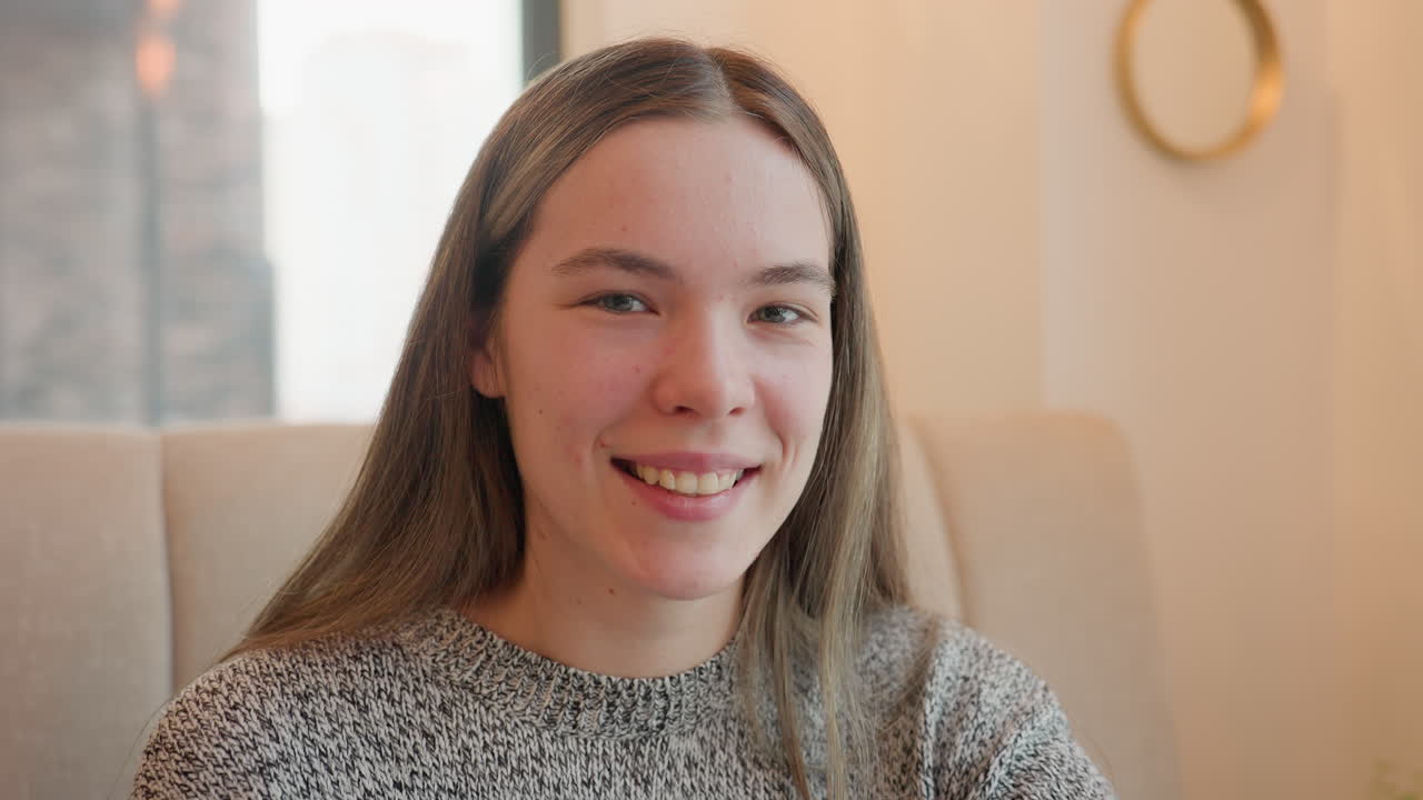 Beautiful young woman seated in brown chair inside tea shop smiling brightly with teeth showing, enjoying relaxing atmosphere with warm lighting, feeling cheerful and content in casual outfit