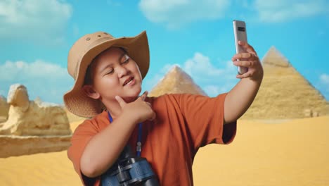asian boy with a hat and binoculars taking a selfie on smartphone while traveling in giza pyramid. boy researcher examines something, travel tourism adventure, close up