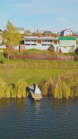 young newlywed couple hugs on little wooden pier by large tranquil river against cottage village on sunny day upper view