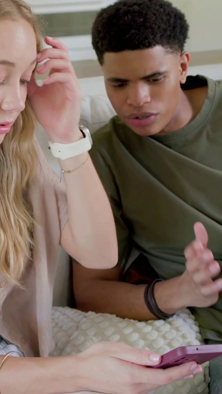 Vertical video: Leaning male in olive tee watching woman handing pink phone on home couch, showing