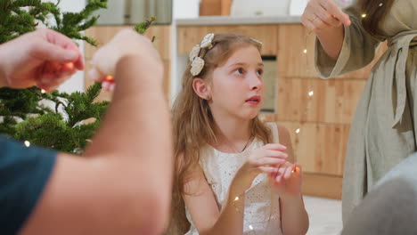 family happily arranging festive lights together, little girl focused and engaged in holiday decoration, cozy home atmosphere with christmas tree in the background