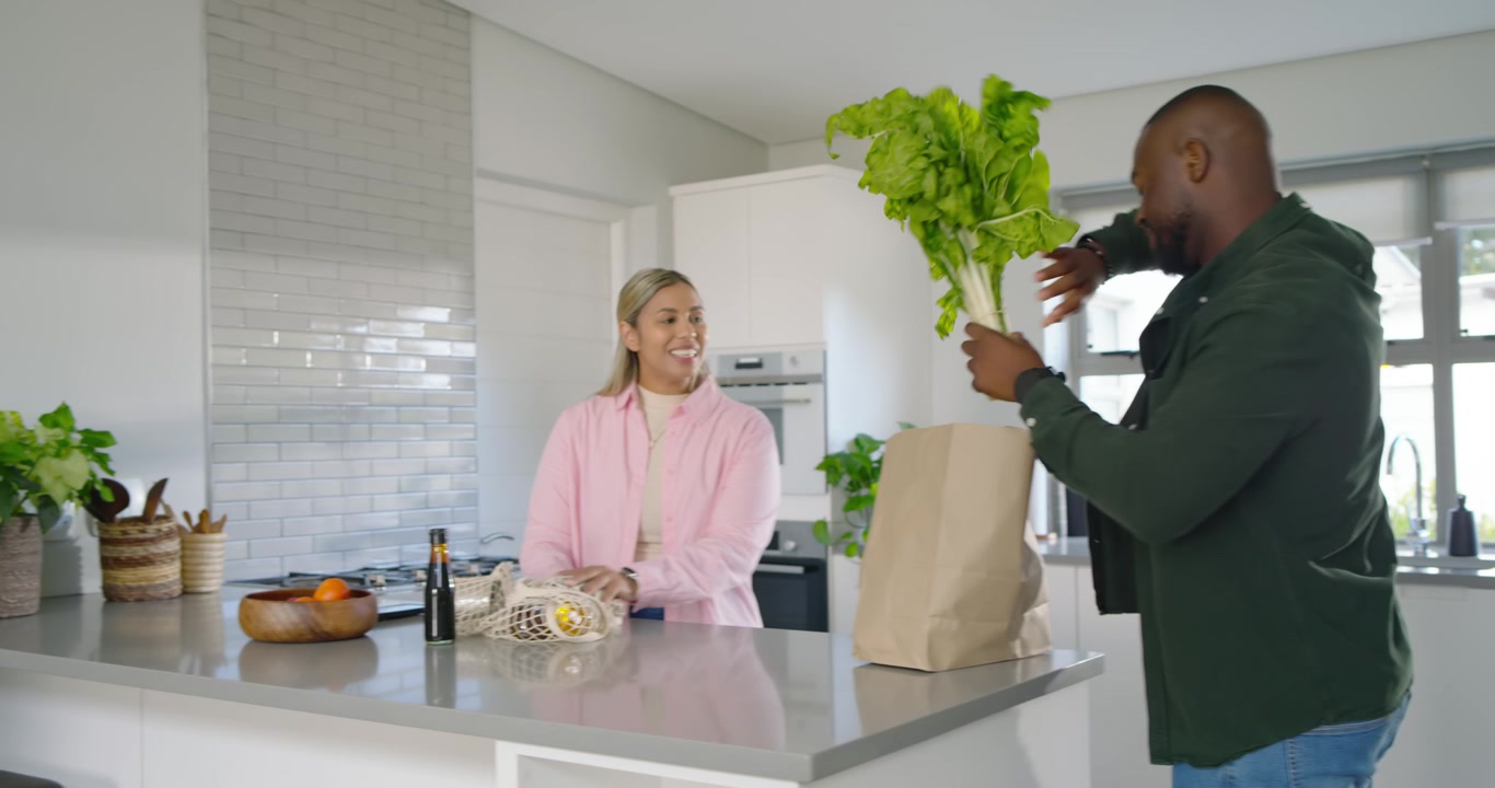 Casal afro-americano organizando as compras na ilha da cozinha, o homem entrando com aipo, guardando alimentos.