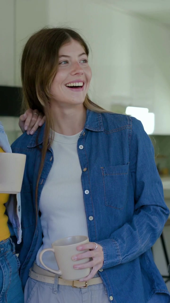 Video verticale: Donna sorridente con top di jeans che tiene una tazza dopo che un'amica le ha messo un braccio sulla spalla sulla porta.