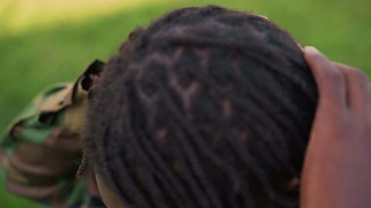 Veteran Calmly Styles Hair Outdoors, Experienced Individual Tidies Dreadlocks Near Green Camp Site, Calm Seasoned Veteran Carefully Arranges Dreadlocks Beside Grassy Area Before Patrol