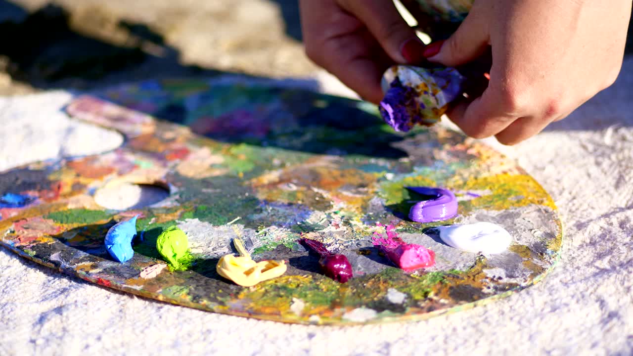 Primo piano, mani femminili con manicure rosso brillante. Tavolozza d'arte. L'artista spreme un po' di vernice viola da un tubetto. C'è un processo creativo. L'artista dipinge all'aperto.