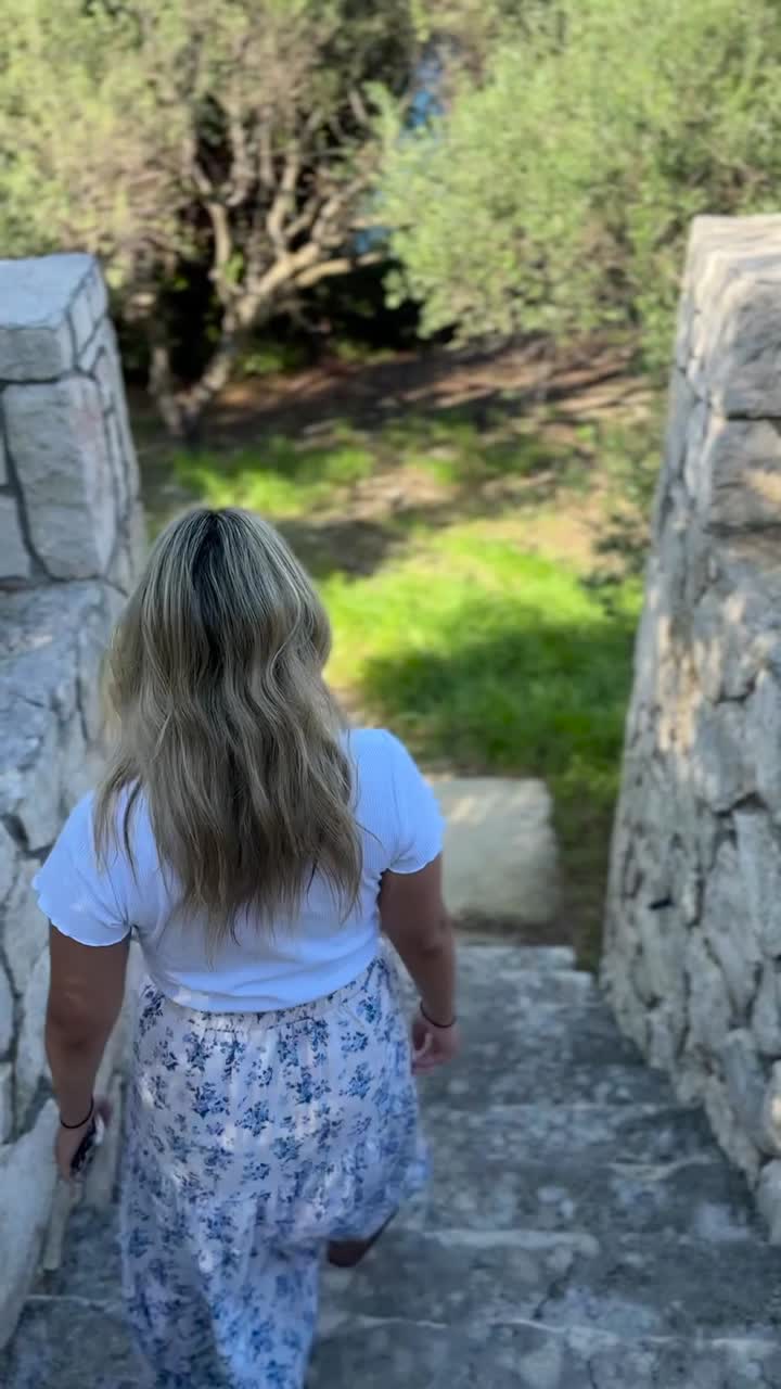 A girl in a long floral skirt walks down a Cycladic-style stone pathway surrounded by lush olive trees in Zakynthos, Greece DOLLY FORWARD