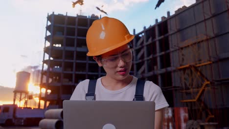 close up de una trabajadora asiática que usa gafas de protección y casco de seguridad usando una computadora portátil y que tiene dolor de cabeza mientras está de pie en el sitio de construcción