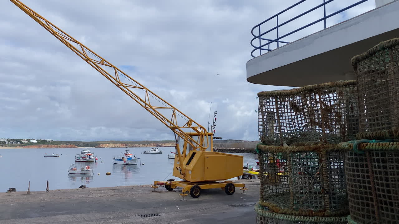 Yellow harbor crane stands beside the calm fishing port of Sagres in the Algarve, with small boats floating on still water under a dramatic cloudy sky