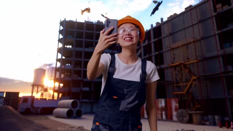 Asian Woman Worker Wearing Goggles And Safety Helmet Waving Hand Having A Video Call On Smartphone While Standing At Construction Site