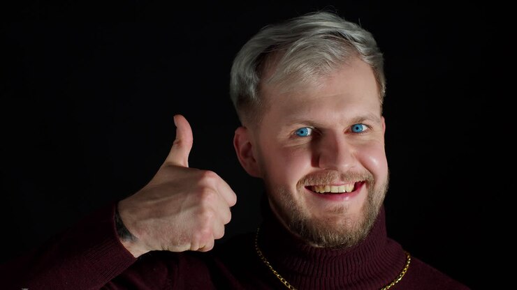 Uomo sorridente con una blusa elegante che guarda con approvazione la telecamera mostrando i pollici su uno sfondo nero.