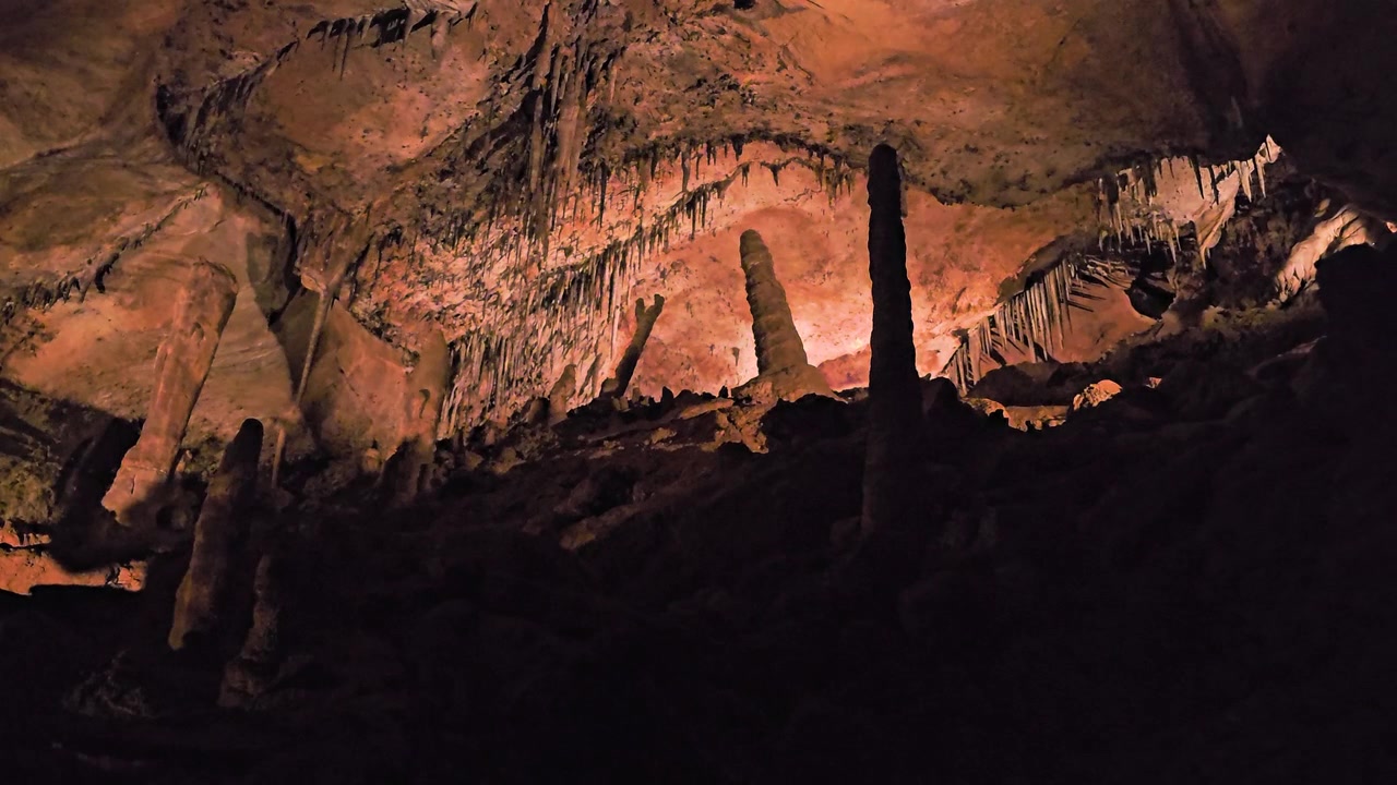 Low angle view inside a cave chamber, showing tall stalagmites, hanging stalactites, pale limestone rocks, dark ceiling textures, and moody underground lighting.