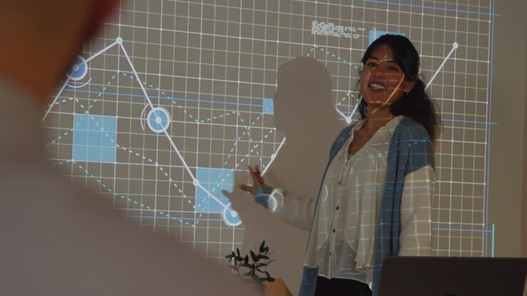 Female Teacher Giving Maths Or Science Lesson In College Or University Standing At The Front Of Class With Projection Of Graph 3