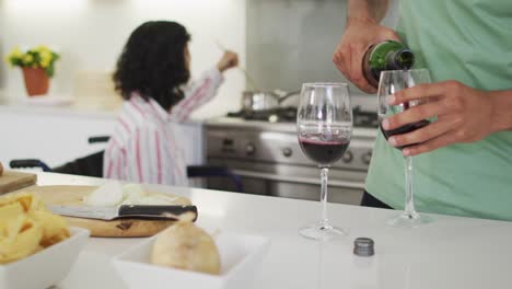 happy biracial woman in wheelchair preparing food and drinking wine with male partner in kitchen