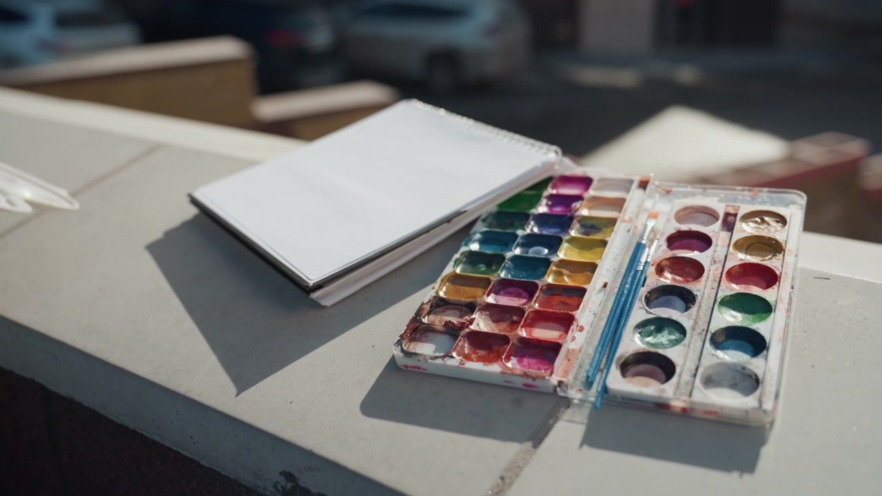 Hände blättern Skizzenbuch über Aquarellpalette um, Straßenstudie im Gange, fokussierte Gesten und nasse Pigmente treffen auf leere Seiten, warmes Licht und sanfter Hintergrundverkehr schaffen eine lebendige Stimmung.
