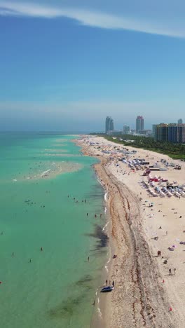 vertical 4k aerial view of miami beach, florida, drone glides above the coastline, capturing a shore full of beachgoers, colorful umbrellas, sunbeds, and turquoise waters under a tropical blue sky.