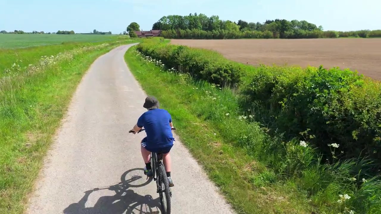 Et barn cykler gennem grønne marker på en solrig dag. Et fugleperspektiv viser den snoede vej omgivet af landskabet. Sollyset kaster lange skygger på grusstien. Cyklisten virker afslappet.