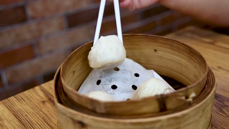 a hand uses chopsticks to pick up a soup dumpling from a bamboo steamer basket on a wooden table, with warm ambient lighting and a brick wall background