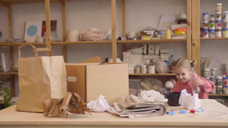 menina loira colocando garrafas plásticas em caixa de papelão sobre uma mesa em uma oficina de artesanato