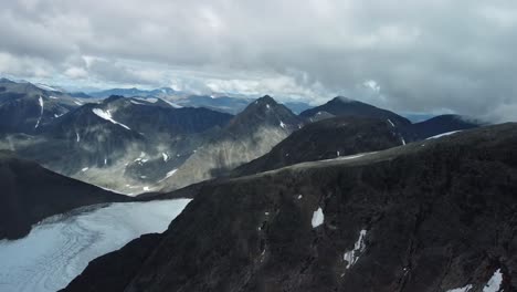 aerial drone footage flying over tall black and dark colored mountains in sweden called kebnekaise hills that have white snow patches and glaciers between them while sun is shining and creates patches