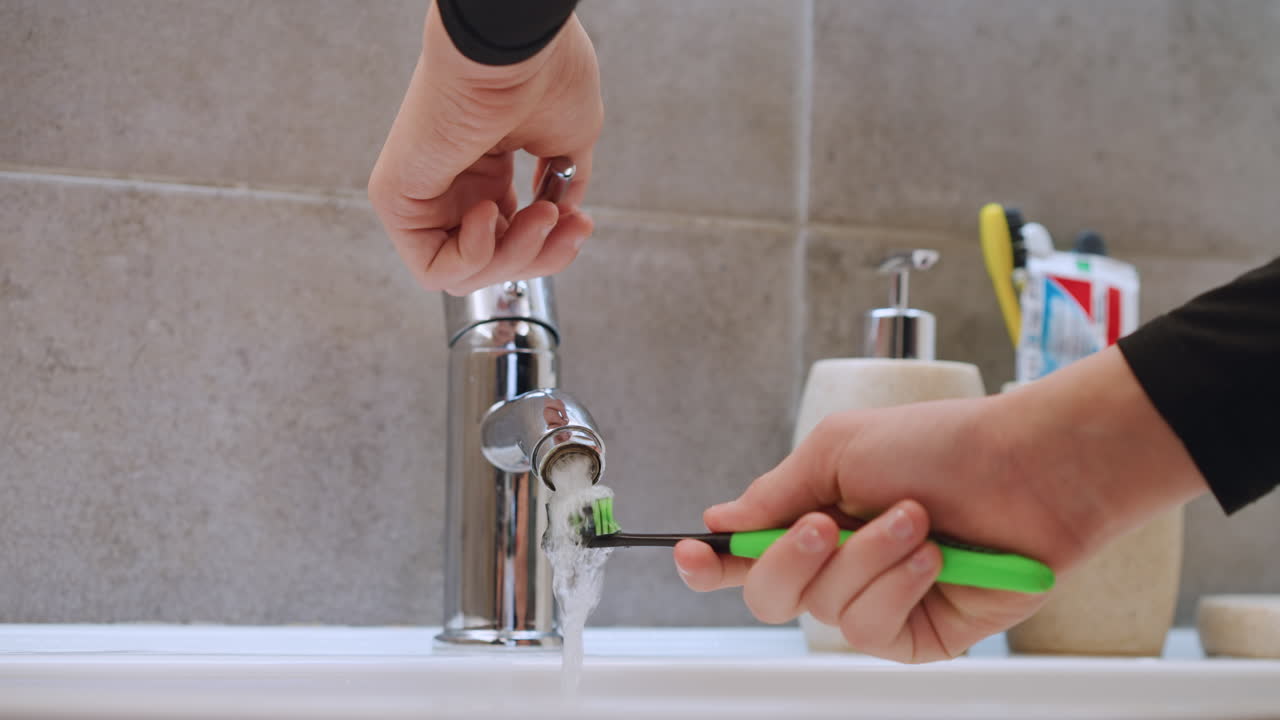 Closeup sink view testing toothbrush under running water, hand turning chrome faucet, ceramic holders and colorful toothbrushes in background, careful motion to rinse bristles, soft neutral tile