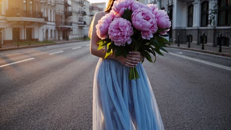 woman walking down urban street during sunset, wearing flowing blue dress and carrying oversized pink peony bouquet partially covering facial features