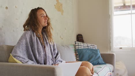 Thoughtful-caucasian-woman-wearing-blanket-sitting-on-sofa-using-laptop-in-sunny-cottage-living-room