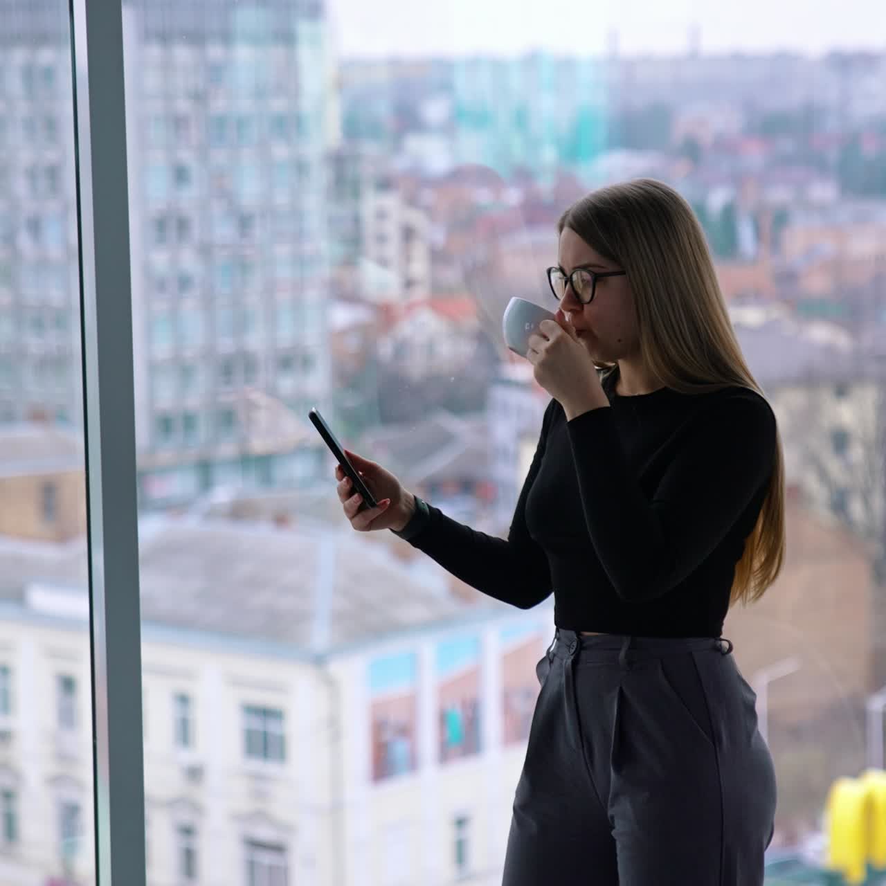 Pretty woman drinks tea and looks at the phone in office. Portrait of a businesswoman in glasses with a cup of coffee standing in the business center at a large panoramic window with city skyscrapers
