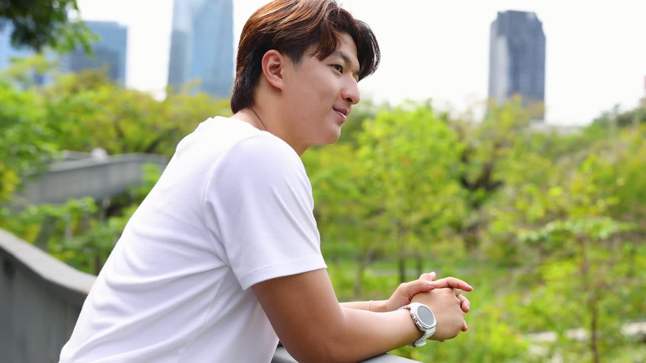 白いTシャツを着た若いタイ人男性が、公園の手すりにもたれかかって微笑んでいます。