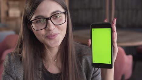 adorable joven mujer de negocios con anteojos está sentada en un café, sosteniendo un teléfono, sonriendo y demostrando una pantalla verde del teléfono