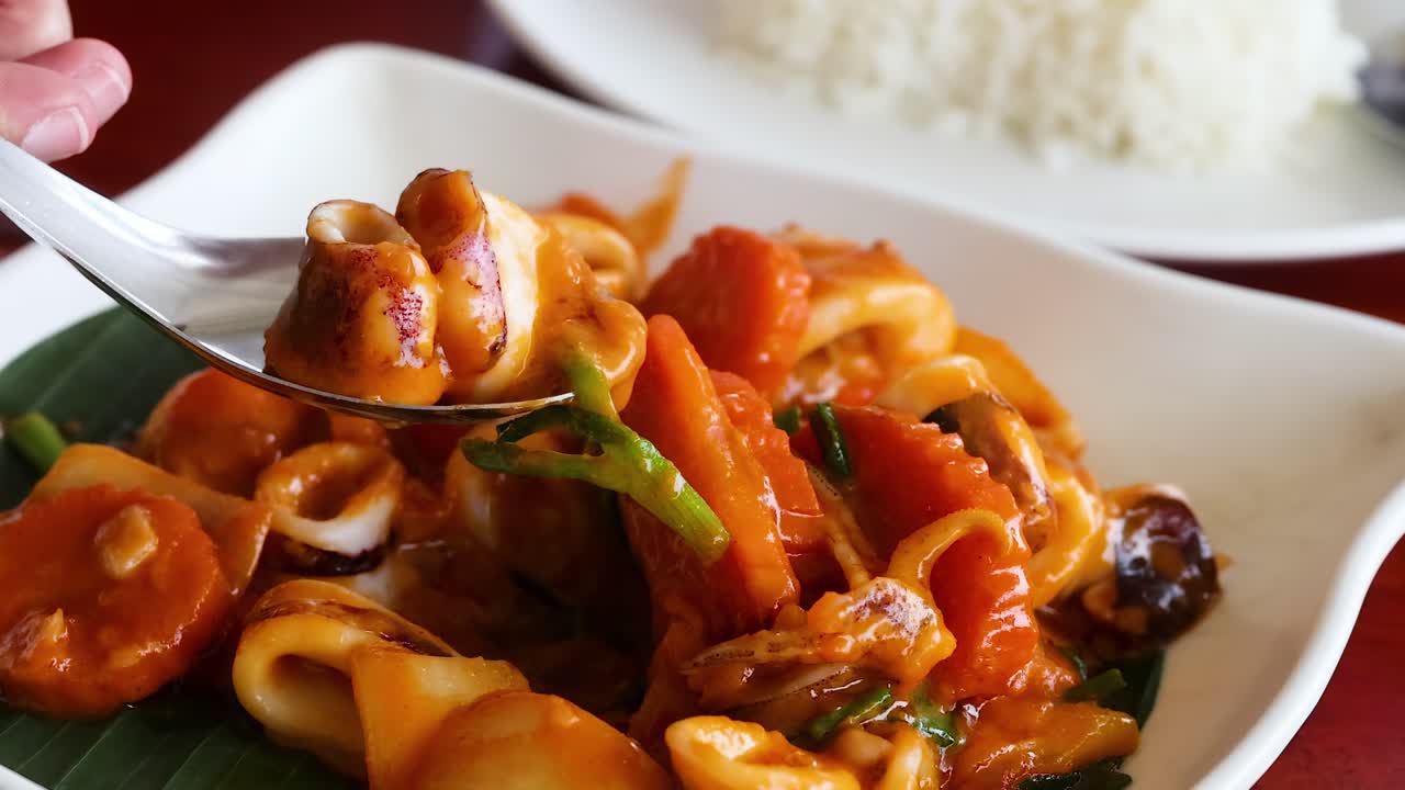 Close-up of a spoon lifting stir-fried squid with carrots and sauce, served with rice.