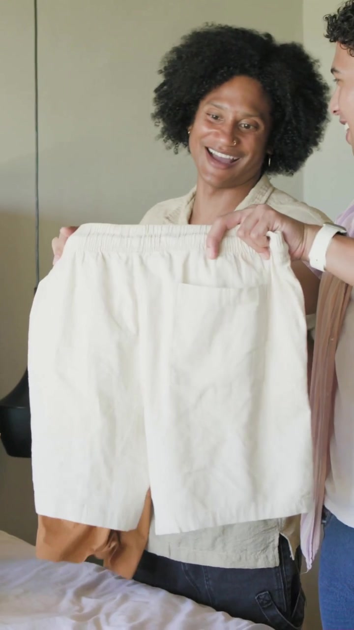Vídeo vertical: Embalando doações, dois homens de camisa bege dobrando roupas na cama para colocar em uma caixa de doação.