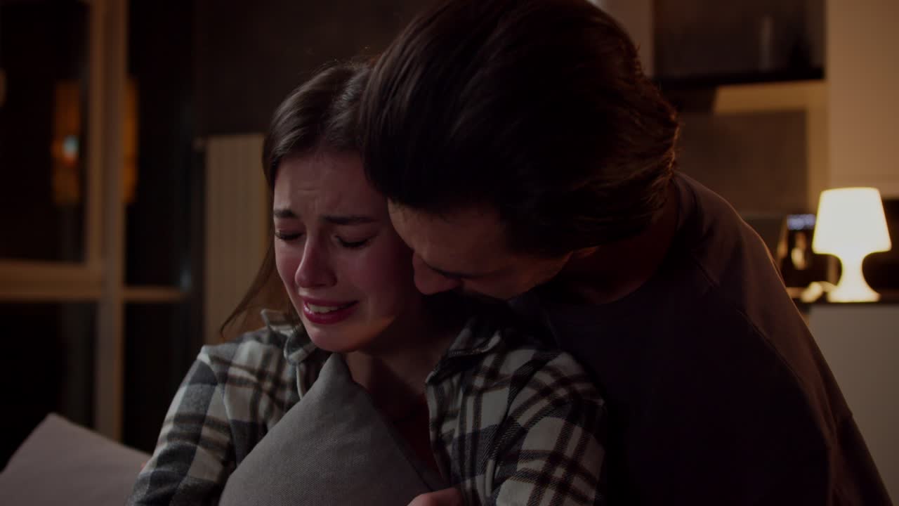 카레드 셔츠를 입은 슬픈 갈색 소녀가 소파에 앉아 울고 베개를 안고 갈색 티셔츠에 그녀의 갈색 남자친구는 저녁에 현대적인 스튜디오 아파트에서 소녀를 지지하고 안습니다.