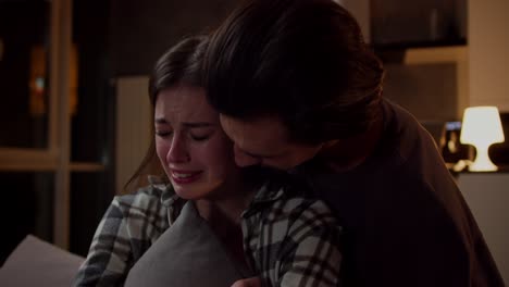 una chica morena triste en una camisa a cuadros se sienta en el sofá llorando y abraza una almohada y su novio moreno en una camiseta marrón apoya y abraza a la chica en un apartamento estudio moderno por la noche