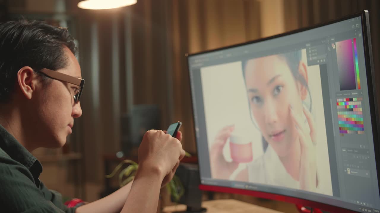 Asian Male Digital Editor Using Mobile Phone While Works In Photo Editing Software On His Personal Computer With Big Display, He Works At Night