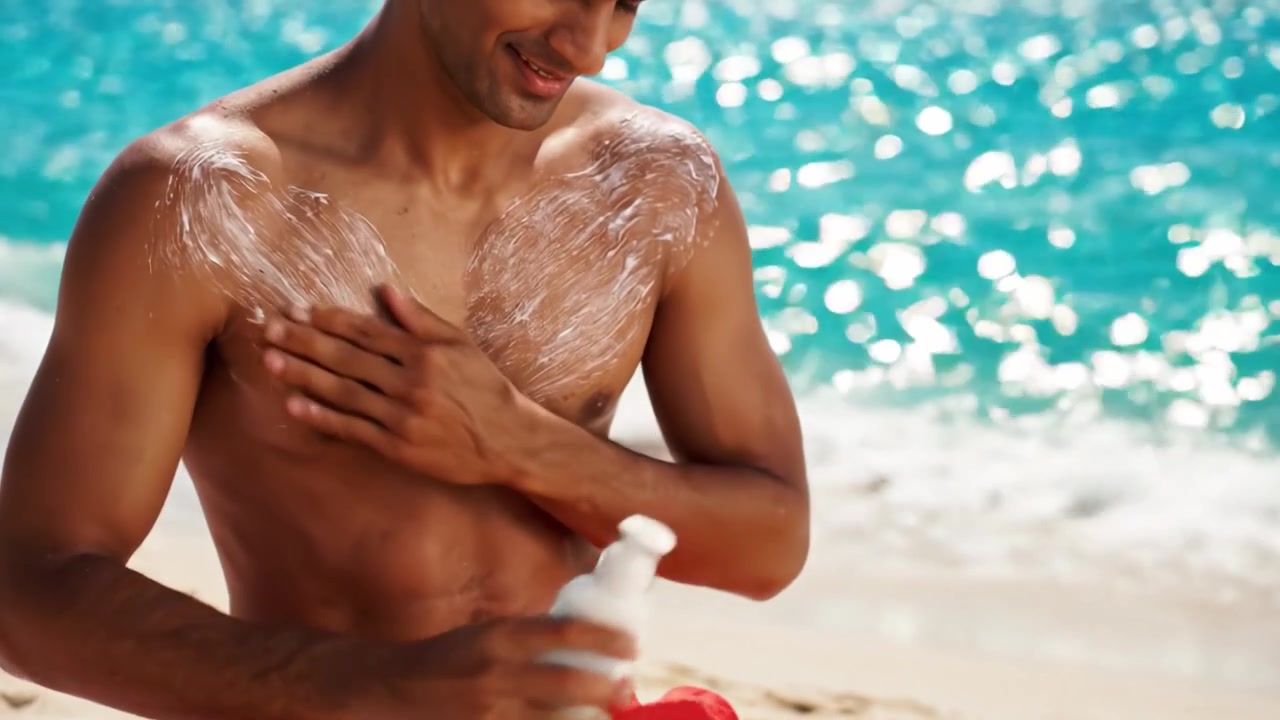Enjoying a Sunlit Beach Moment: A Young Man Applies Sunscreen to Protect His Skin from Harmful UV Rays While Relaxing by the Sparkling Ocean Waves