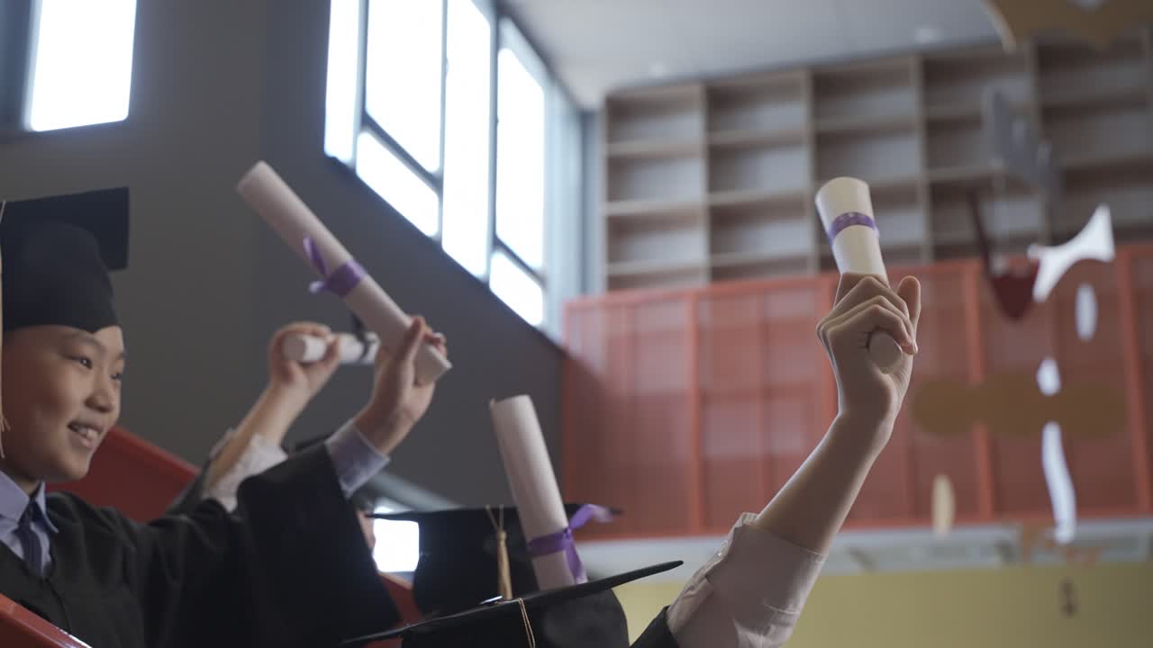 grupo de felices estudiantes de preescolar multirraciales con toga y birrete parados en las escaleras y mostrando sus diplomas de graduación