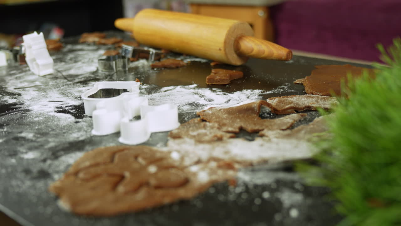 Star, tree and gingerbread man shapes are cut into rolled dough on a floured counter with cookie cutters and a rolling pin, festive home kitchen atmosphere, real time