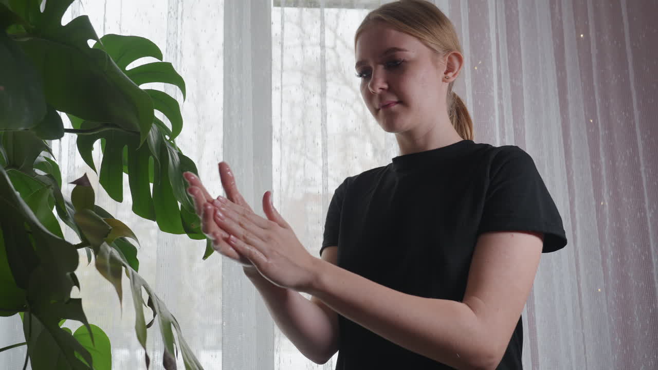 Light skin woman in black shirt gently rubbing oily palms together near green leaf and sheer white curtain, calm indoor ambiance with natural daylight and focused on self-care and relaxation
