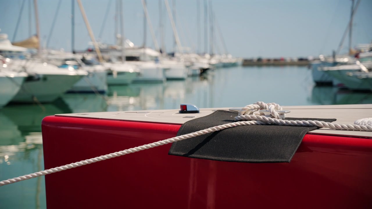Primo piano cinematografico di una barca a vela rossa ormeggiata in un porto turistico con yacht sfocati sullo sfondo.