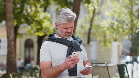 Happy-rich-mature-man-counting-holding-money-dollar-cash-use-smartphone-calculator-app-booking-hotel