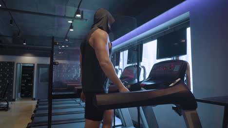 unrecognizable sporty man with muscular arms walking on treadmill inside gym in slow motion, wearing hood covering face - profile view