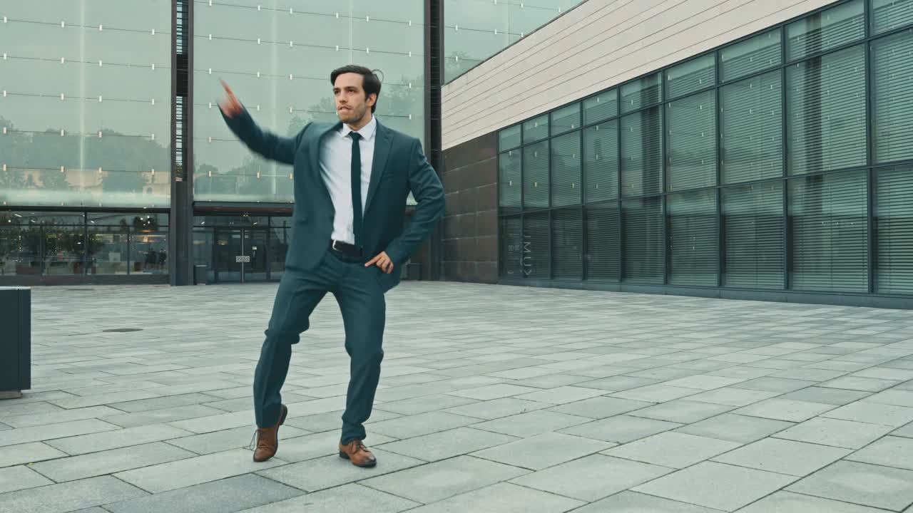 un hombre de negocios alegre y feliz en un traje está bailando activamente en una plaza de la calle. escena rodada en un parque de hormigón urbano al lado del centro de negocios. día soleado.