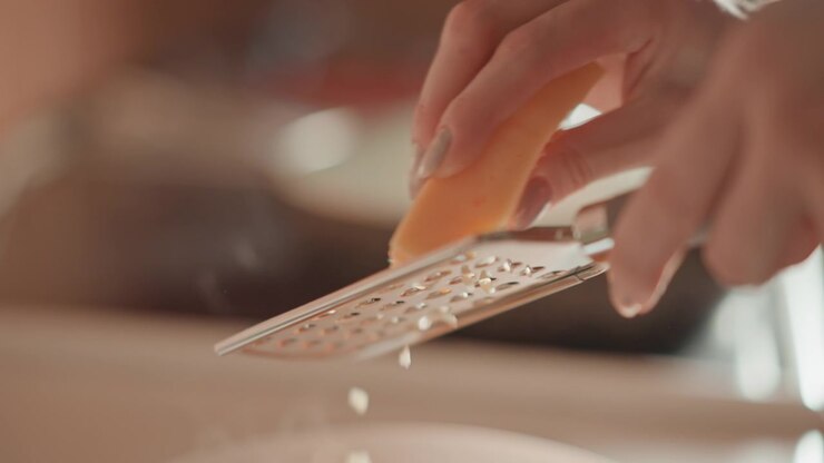 Tight low-angle view shows hands rubbing cheese block against handheld metal grater, turning solid cheese into thin shavings and small fragments that fall below for home cooking, garnish, and meal