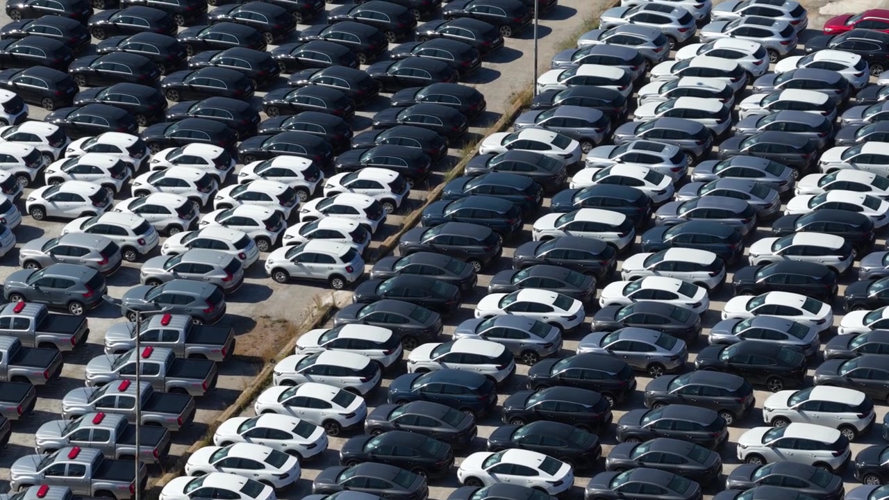 Luftfoto ovenfra af en kæmpe parkeringsplads med hundredvis af nye biler perfekt arrangeret i rækker, der venter på distribution og salg fra bilfabrikkens lagerområde.