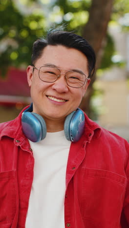 Portrait-of-happy-smiling-Asian-man-listening-music-taking-off-headphones-looking-at-camera-outdoors