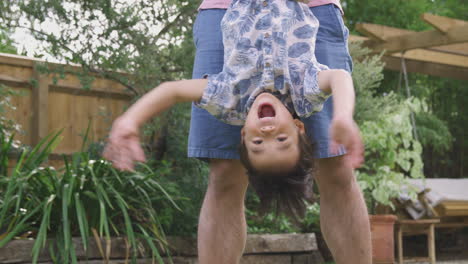 asian father holding son upside down by legs in garden as they play game together