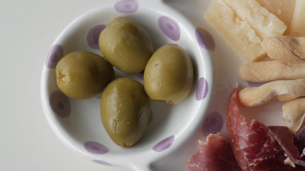 Four green olives in a white bowl with purple dots alongside cheese and jamón